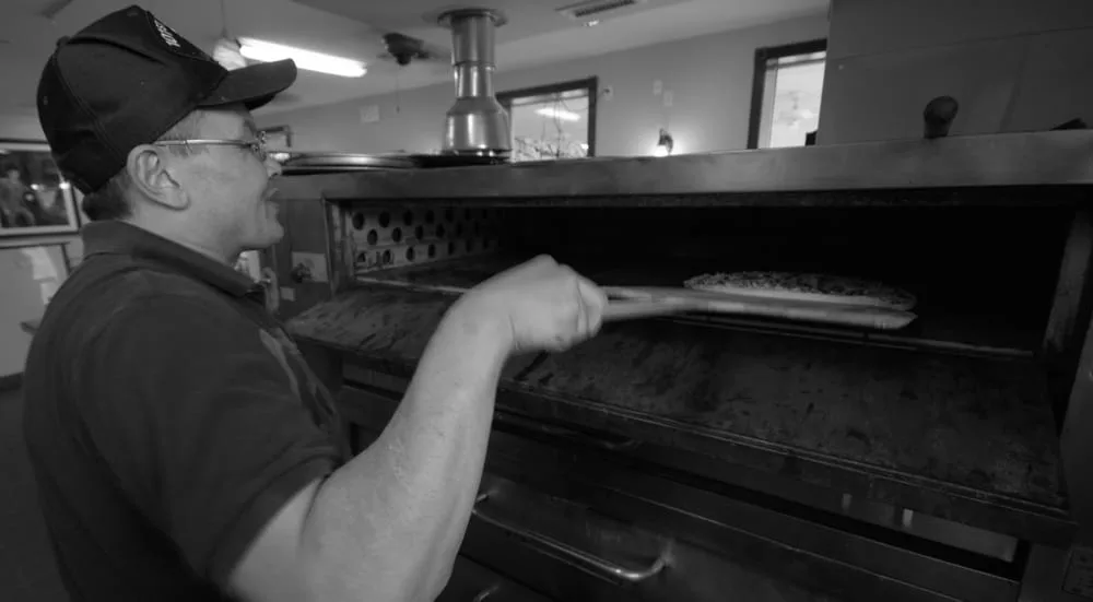 Black and white photo of a cook sliding a pizza into the brick oven on a wooden peel