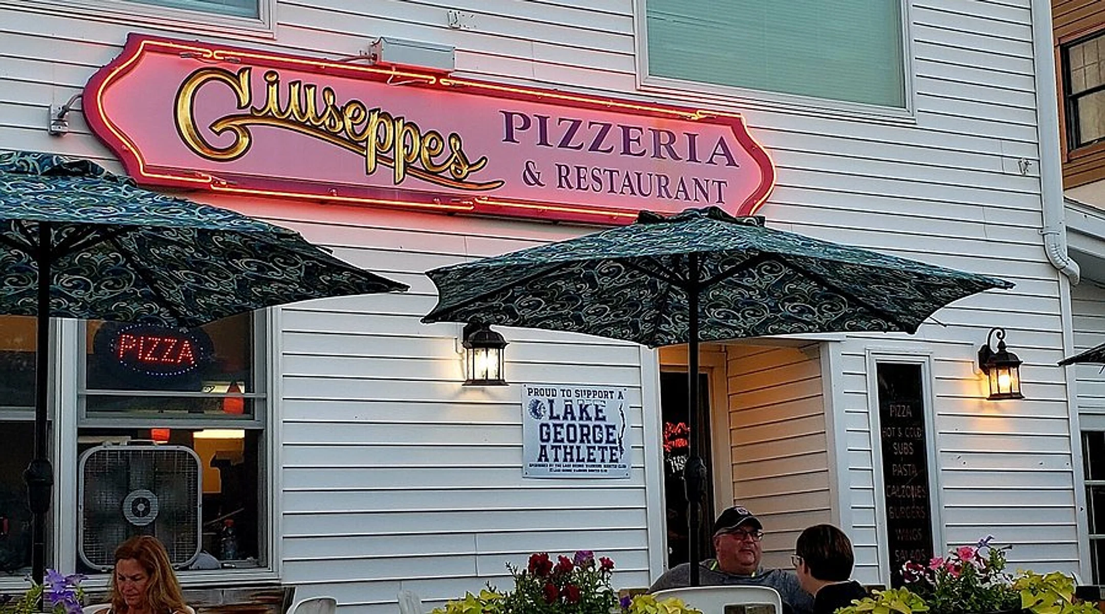 Giuseppe's Pizzeria & Restaurant storefront straight-on at dusk, neon sign glowing above the patio, Lake George, NY